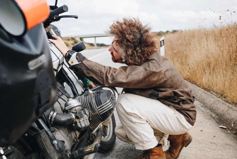 man in white pants and a leather jacket sits and looks at his bike on the kerb